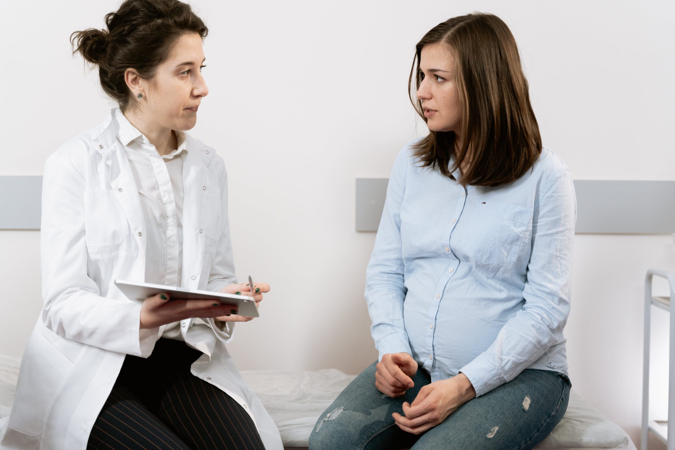 light skinned doctor with dark hair is holding a clipboard and speaking with a fair skinned woman with dark hair who is pregnant