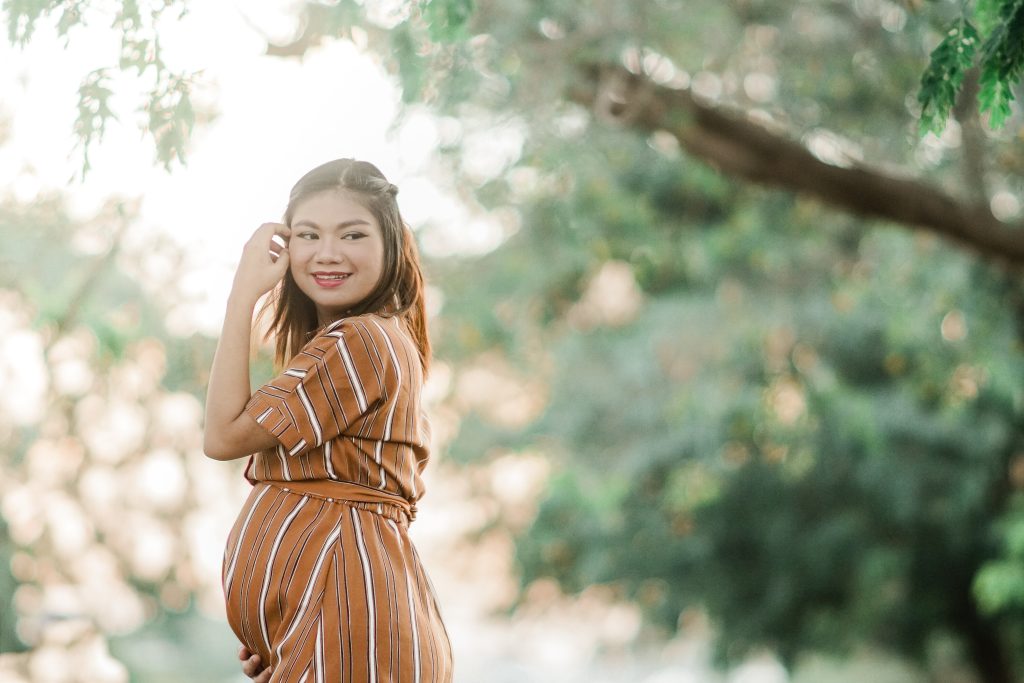 Brown haired pregnant woman in a striped orange dress standing outside, pushing hair behind her ear