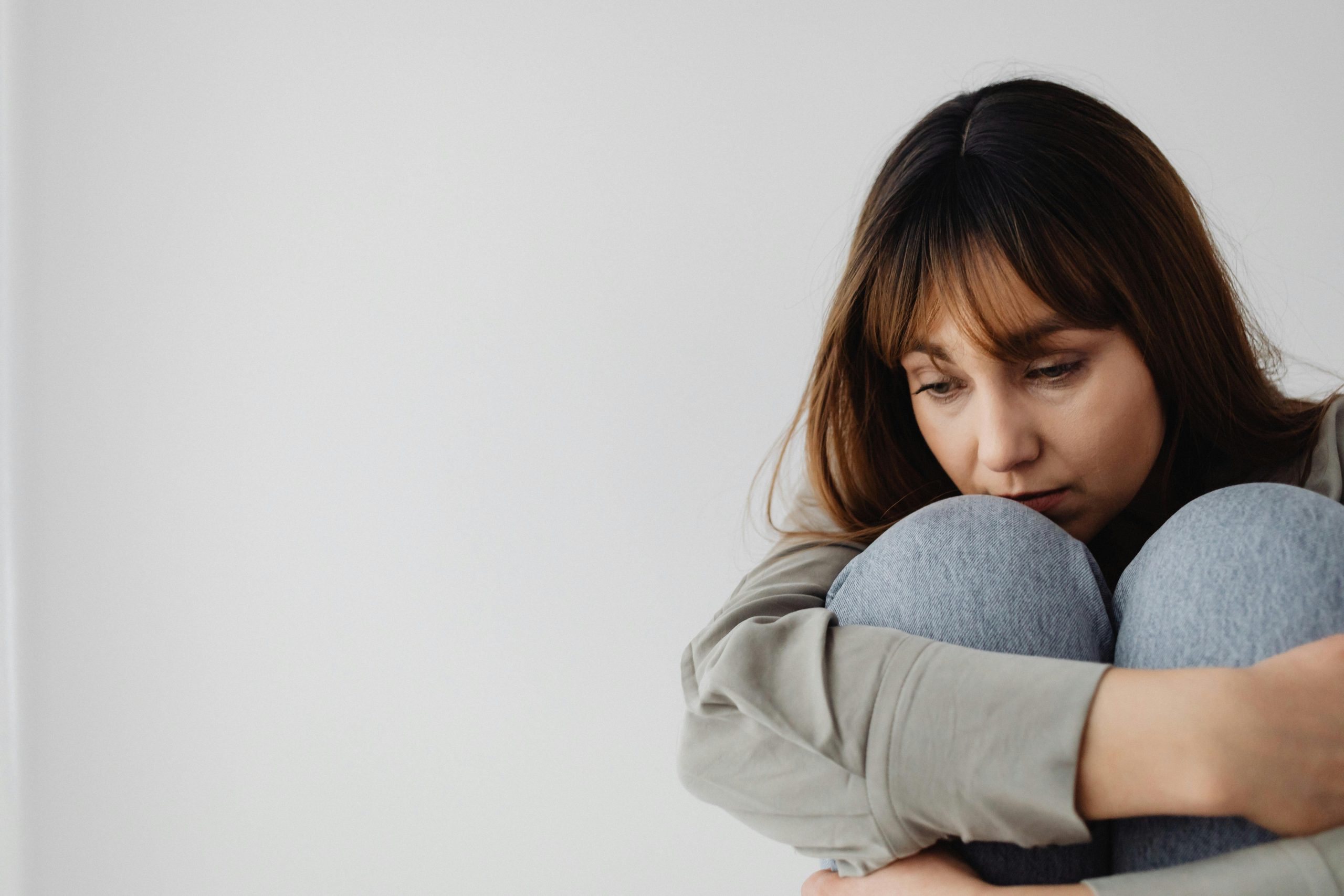 on the right side of the image, a woman with brown hair in a gray long sleeve shirt and light blue jeans hugging her knees, looking down in anxiety