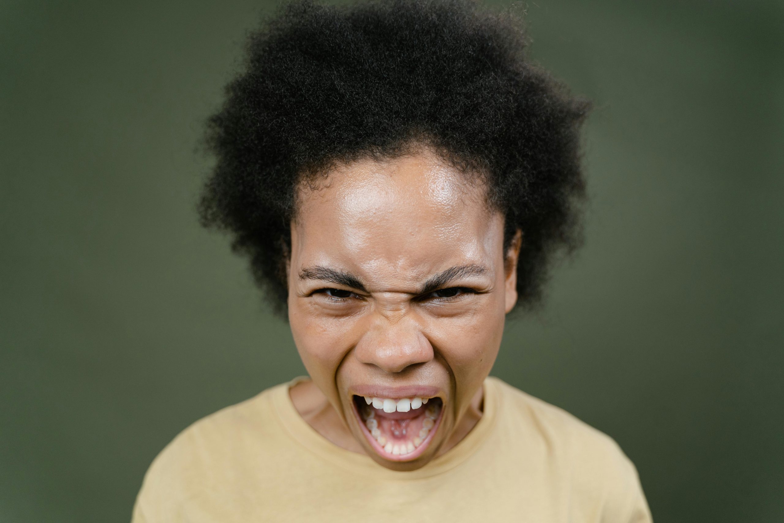Brown skinned woman in yellow shirt and tight, short curly. black hair is screaming in anger against a breen backdrop